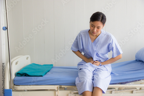 Asian young woman in patient's dress was touching her stomach and sitting on the bed in the hospital's patient room with Saline bottle . She has gotten a severe stomachache due to food poisoning