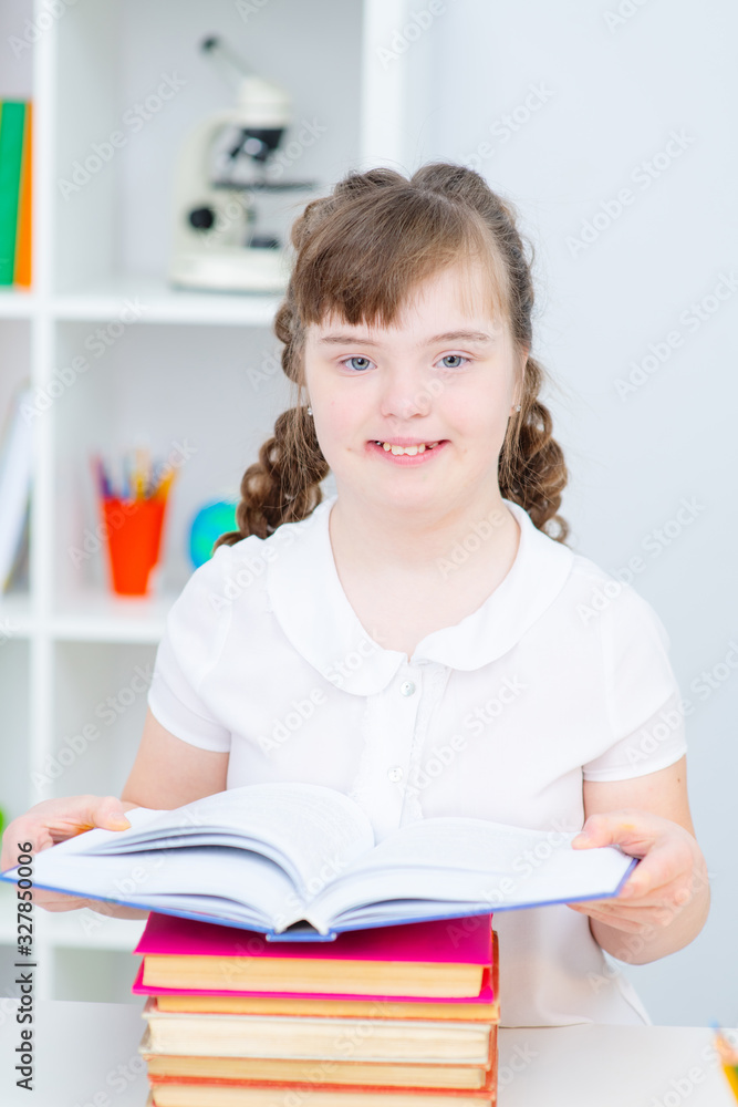 A girl with down syndrome sitting at home at the table reading an open book lying in front of her. Girl with Down Syndrome looking thoughtfully to the side A girl with down syndrome sitting at home at the table reading an open book lying in front of her. Girl with Down Syndrome looking thoughtfully to the side