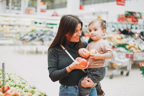 Young woman mother with cute baby boy toddler child on hands buys the fresh apples in supermarket