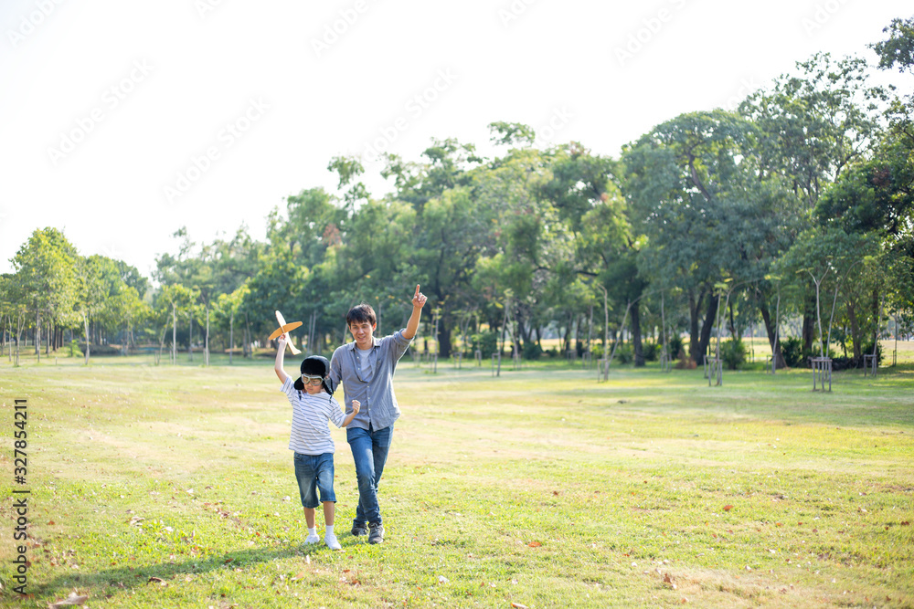 Happy Asian father playing paper plane model with son at park, boy wearing pilot helmet, holding airplane toy in the air, Dad looking proud and support to his kid dream goal, family insurance concept