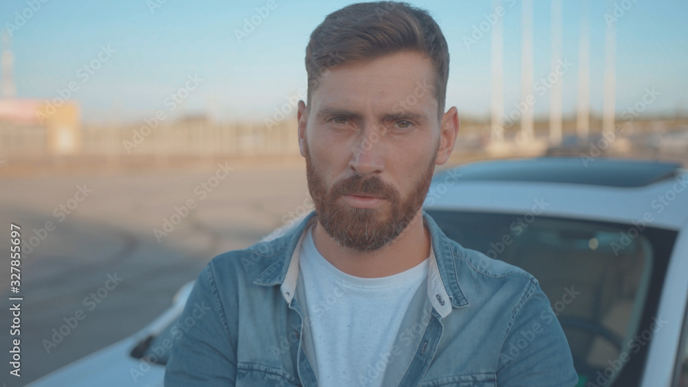 Portrait of severe confident guy leaning on car at parking space. Close-up handsome serious young man driver against modern auto under sunset. Portrait of severe confident guy leaning on car at parking space. Close-up handsome serious young man driver against modern auto under sunset.