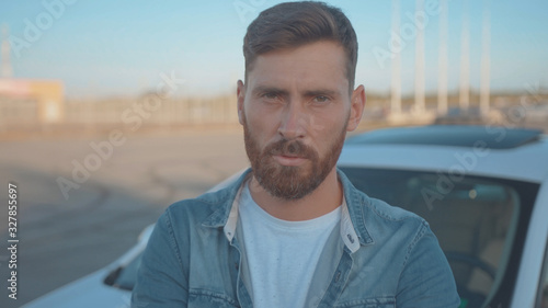 Portrait of severe confident guy leaning on car at parking space. Close-up handsome serious young man driver against modern auto under sunset.