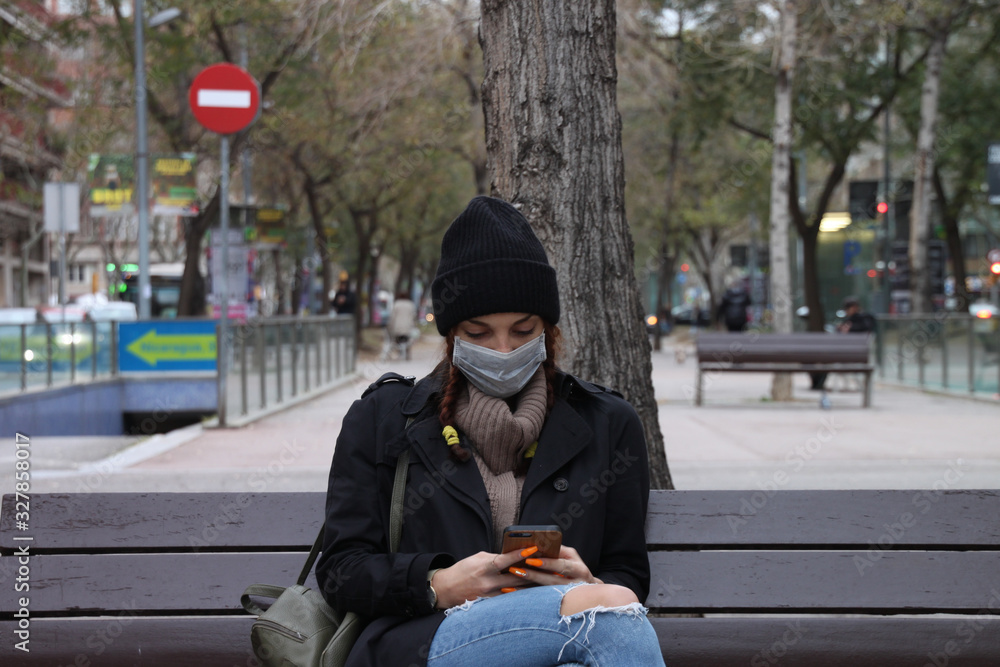 Obraz premium Young woman sitting on a street bench wearing medical mask and looking at her smartphone