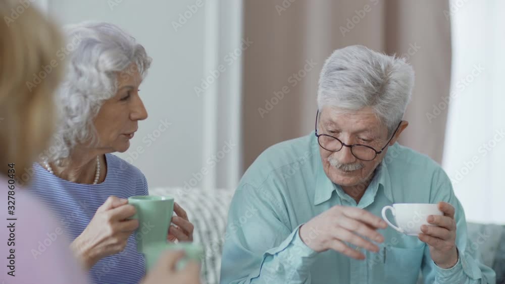 Panning medium shot of group of senior friends talking over tea at home. Two elderly women listening to man in eyeglasses Panning medium shot of group of senior friends talking over tea at home. Two elderly women listening to man in eyeglasses