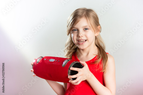 child 7 - 8 years old, cute charming blonde girl with blond hair in a red dress with a red bluetooth speaker listens to music and smiles, gray background