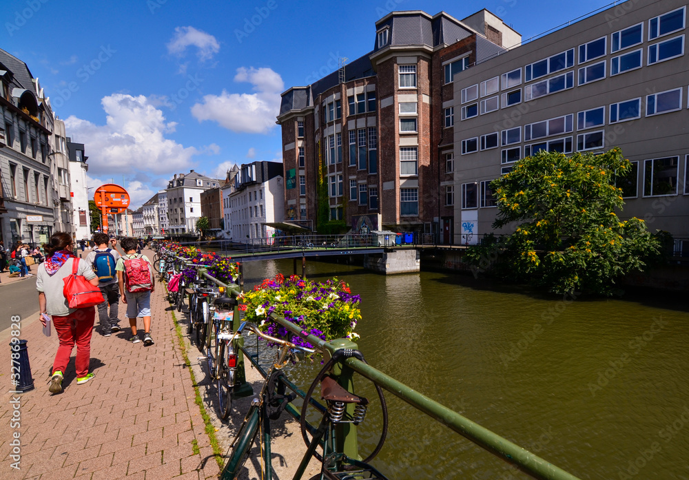 Ghent, Belgium, August 2019. Planters with brightly colored plants and some parked bikes stand out along the canals. People walk along the sidewalk that runs alongside the water.