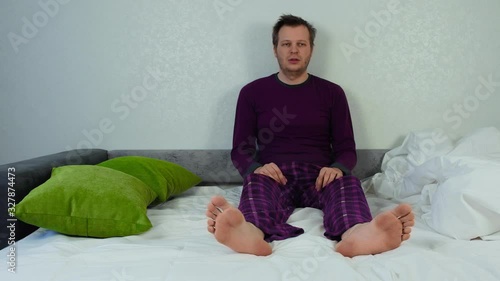 Confused and sleepy man is sitting on a bed. Depression concept, doldrums. Problems in the family and at work. Caucasian unshaven and unkempt guy in pajamas. Wide shot slider movement, tracking right