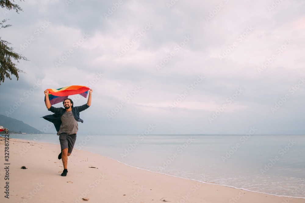 Cheerful guy with a rainbow flag on the beach.