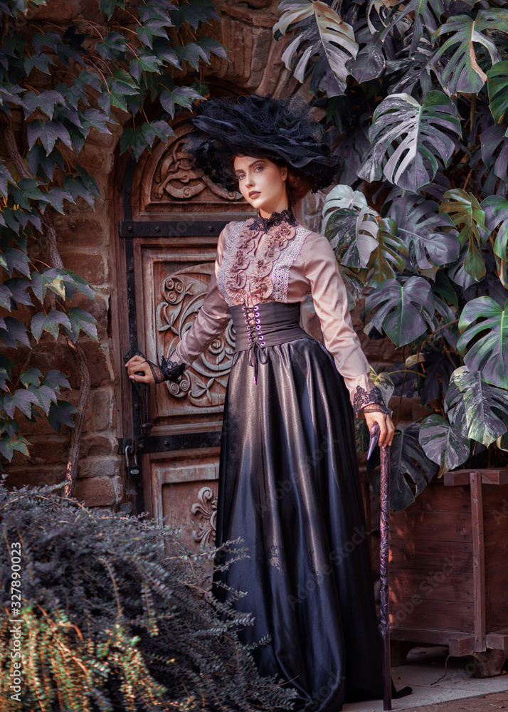 Portrait of amazing cute young woman in big lacy black hat and vintage brown blouse with black long flowing skirt stands near wooden carved antique door in green thicket and looks at camera