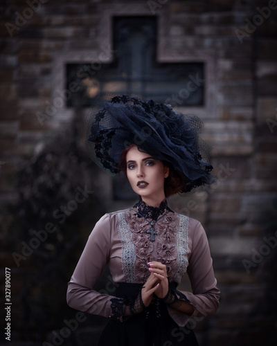 Close up portrait of amazing cute young woman in large lacy black hat and vintage brown blouse stands and looks at camera on background dark brick wall