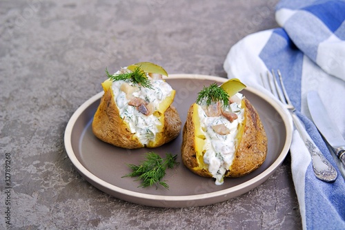Whole baked potatoes with sour cream, herring and pickled cucumbers sauce on a brown plate on a gray concrete background. Potato recipes. Copyspace.