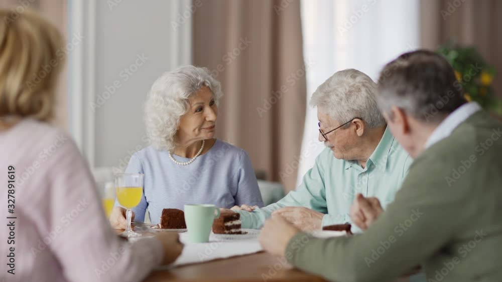 Group of four senior friends sitting at dining table at home and enjoying talk over dessert. Smiling elderly woman with curly grey hair speaking Group of four senior friends sitting at dining table at home and enjoying talk over dessert. Smiling elderly woman with curly grey hair speaking