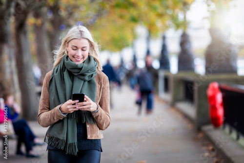 Eine attraktive, blonde Frau läuft auf der Straße und liest ihre Emails und Nachrichten auf dem Mobiltelefon photo