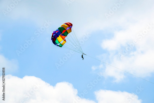 Multi-colored parachute with man soars in the blue sky between white clouds.