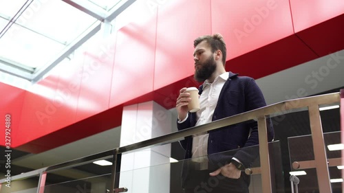 Young bearded man is drinking take-out coffee, standing near glass fence in contemporary business center. Close-up view of calm brunette guy with cup of the coffee to go in modern building with red
