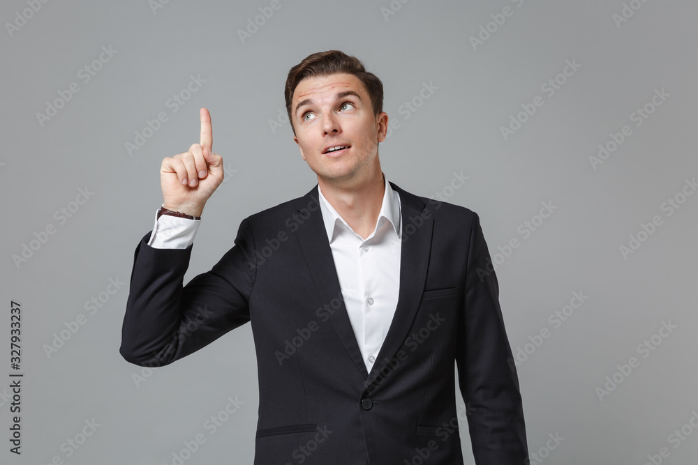 Pensive young business man in classic black suit shirt posing isolated on grey background. Achievement career wealth business concept. Mock up copy space. Holding index finger up with great new idea. Pensive young business man in classic black suit shirt posing isolated on grey background. Achievement career wealth business concept. Mock up copy space. Holding index finger up with great new idea.