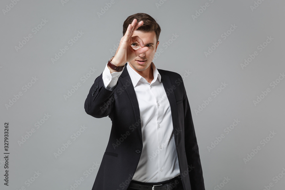 Young business man in classic black suit shirt isolated on grey background. Achievement career wealth business concept. Mock up copy space. Holding hand near eye, imitating eyeglasses or binoculars. Young business man in classic black suit shirt isolated on grey background. Achievement career wealth business concept. Mock up copy space. Holding hand near eye, imitating eyeglasses or binoculars.