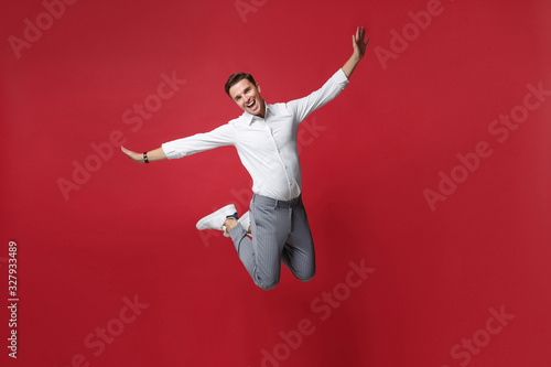 Funny young business man in white shirt, gray pants posing isolated on bright red background studio portrait. Achievement career wealth business concept. Mock up copy space. Jumping, spreading hands.