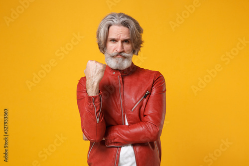 Angry concerned elderly gray-haired mustache bearded man in red leather jacket posing isolated on yellow orange wall background in studio. People lifestyle concept. Mock up copy space. Clenching fist.