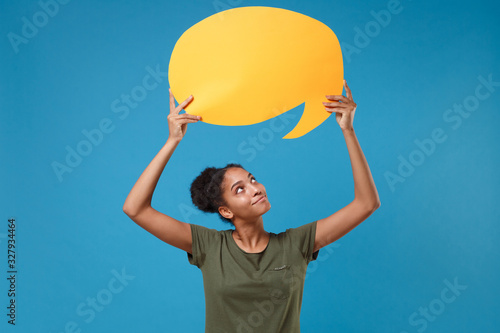 Pretty young african american woman girl in casual t-shirt posing isolated on bright blue background. People lifestyle concept. Mock up copy space. Hold yellow empty blank Say cloud, speech bubble. photo