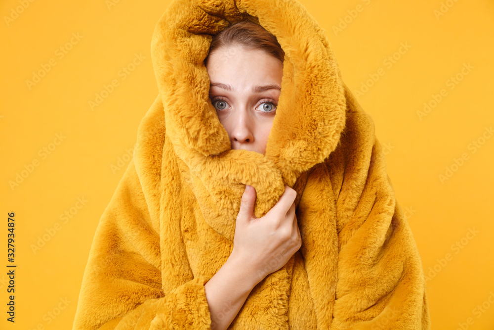 Frozen shocked young blonde woman girl posing isolated on orange wall background studio portrait. People sincere emotions lifestyle concept. Mock up copy space. Wrapping herself in yellow fur coat.