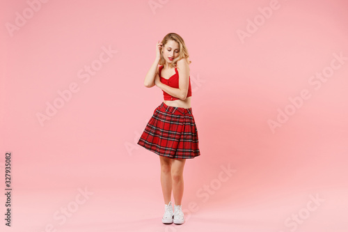 Stunning young blonde woman girl in red sexy clothes isolated on pastel pink wall background studio portrait. People emotions lifestyle concept. Mock up copy space. Looking down holding hands crossed.