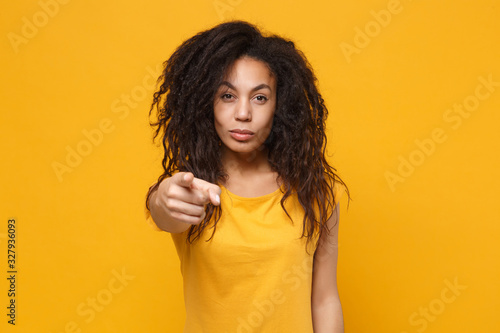 Attractive young african american woman girl in casual t-shirt posing isolated on yellow orange background studio portrait. People lifestyle concept. Mock up copy space. Point index finger on camera.
