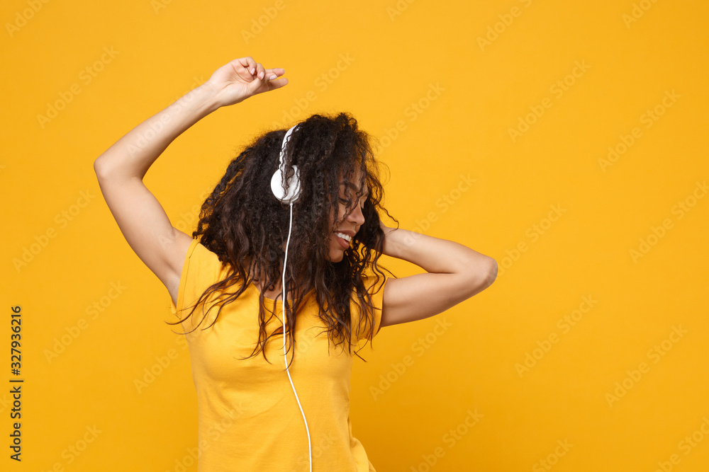 Fototapeta premium Cheerful young african american woman girl in casual t-shirt posing isolated on yellow orange background in studio. People lifestyle concept. Mock up copy space. Listen music with headphones, dancing.