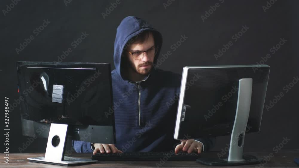 Serious caucasian hacker male sit at table, look at screen of laptop studying computer system and typing password for hacking bank account or online bullying, sit isolated in dark space, black Serious caucasian hacker male sit at table, look at screen of laptop studying computer system and typing password for hacking bank account or online bullying, sit isolated in dark space, black