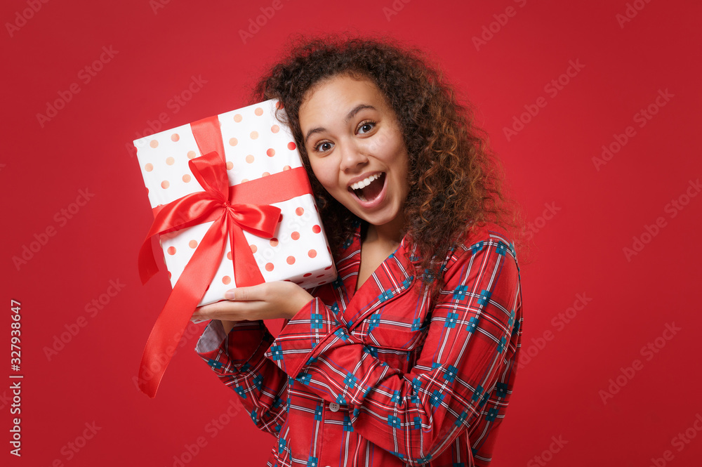 Amazed young african american girl in pajamas homewear posing resting at home isolated on red background. Relax good mood lifestyle concept. Mock up copy space. Hold present box with gift ribbon bow. Amazed young african american girl in pajamas homewear posing resting at home isolated on red background. Relax good mood lifestyle concept. Mock up copy space. Hold present box with gift ribbon bow.