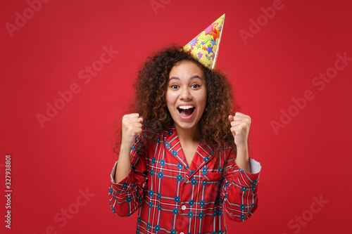 Joyful african american girl in pajamas homewear, birthday hat rest at home isolated on red background. Relax good mood lifestyle concept. Mock up copy space. Keeping mouth open doing winner gesture.
