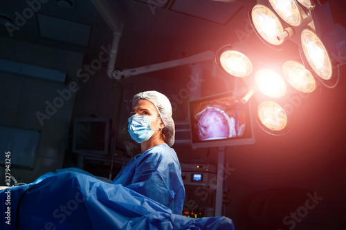 Young woman doctor in cap and face mask in surgery room interior on dark background with lights on.