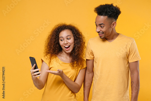 Excited young friends couple african american guy girl in casual clothes isolated on yellow background. People sincere emotions lifestyle concept. Mock up copy space. Pointing hand on mobile phone.