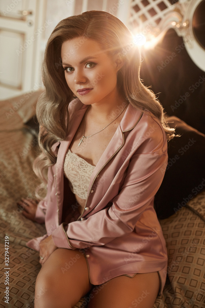 Beautiful young woman with platinum blonde hair in satin pink pajamas sits on the bed. Model girl with perfect skin face and wavy hairstyle posing in a bedroom of a luxury hotel apartment Beautiful young woman with platinum blonde hair in satin pink pajamas sits on the bed. Model girl with perfect skin face and wavy hairstyle posing in a bedroom of a luxury hotel apartment