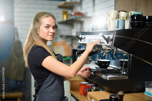 Young girl at work stands by the coffee machine and smiles. Cute blonde girl at work in a coffee shop