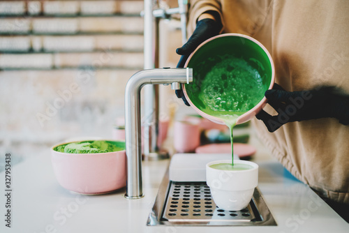 Fototapeta Vegetarian natural matcha tea made by professional barista in asian cafe. Female's hands holding a bowl with a freshly made matcha tea pouring to the white cup. Tea ceremony in a oriental style.