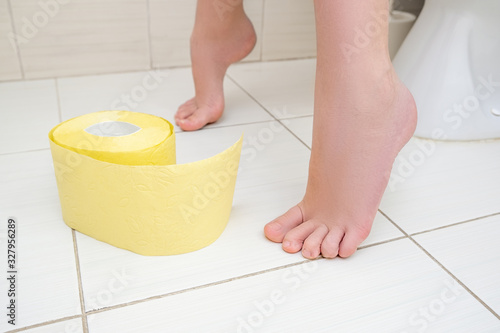 Cropped legs of a boy sitting on a toilet with a roll of yellow toilet paper on a floor, digestive problems and difficulties in toilet concept
