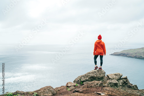 Trail runner female dressed orange sporty hoodie and red cap resting on the cliff and enjoying Atlantic ocean view on Ponta de Sao Louren  o peninsula -the easternmost point of Madeira island  Portugal