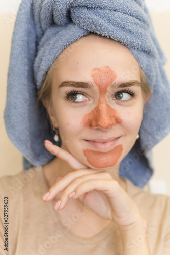 A beautiful girl with a towel on her head makes a multi mask on the t-zone of orange color with a scrub. The cute blonde is having fun doing a home care, moisturizing and smoothing facial mask photo