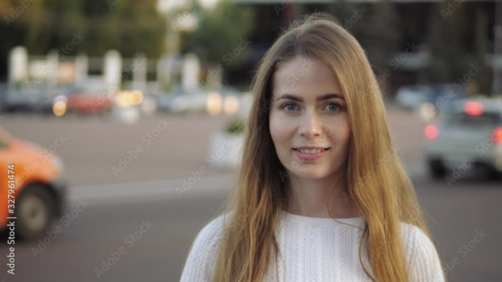 Medium close up slow motion head shot of young adorable blonde elegant woman with blue eyes looking at the camera changing facial expression on city street golden hour warm evening sun moving cars on Medium close up slow motion head shot of young adorable blonde elegant woman with blue eyes looking at the camera changing facial expression on city street golden hour warm evening sun moving cars on