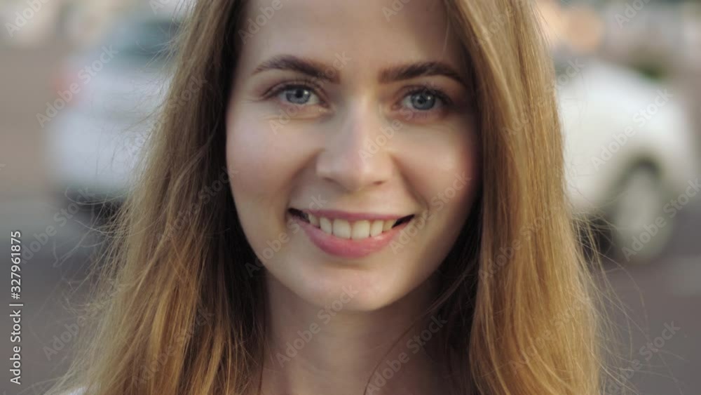 Big close up slow motion head shot of young adorable blonde elegant woman with blue eyes looking at the camera changing facial expression from serious to adorable smile on city street golden hour warm Big close up slow motion head shot of young adorable blonde elegant woman with blue eyes looking at the camera changing facial expression from serious to adorable smile on city street golden hour warm