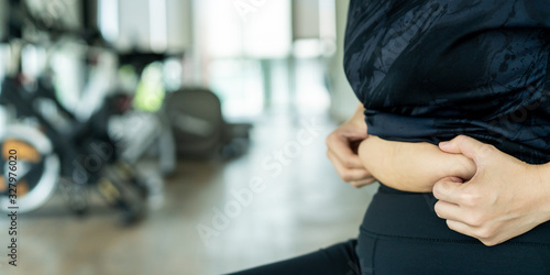 Asian fat woman grabbing on her stomach belly. Fat woman worried about overweight problem and trying the exercise in the gym. Fatness in unhealthy eating people concept. Photograph with copyspace.