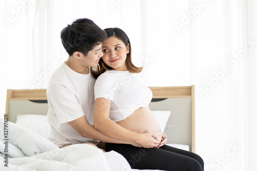 Asian lovely husband and wife portrait, his wife is in pregnancy. Husband tenderly hug and kiss his wife on the bed with care. Husband embracing his pregnant wife and making their hands in heart shape © DG PhotoStock