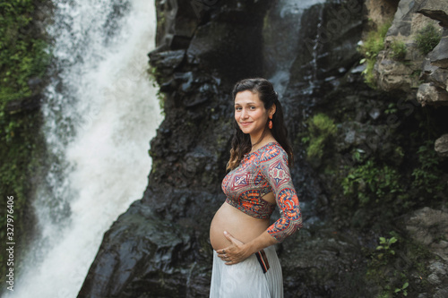 Portrait of young authentic pregnant woman near waterfall outdoors. Smiling and happy  holding belly. Spiritual harmony with nature. Travel and childbirth in Bali.