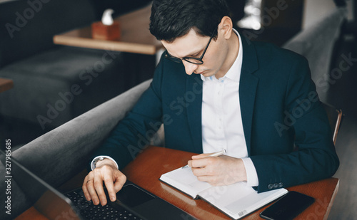 Caucasian worker dressed in a suit is using a laptop while writing something sitting in a restaurant