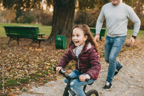 Playful caucasian father walking with his daughter in the park while she is riding a bike happily