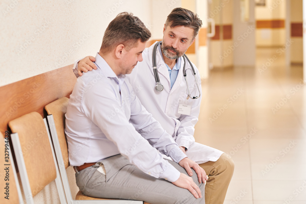 Mature bearded doctor with stethoscope around neck sitting chair in corridor and talking to man worried about relative in surgery Mature bearded doctor with stethoscope around neck sitting chair in corridor and talking to man worried about relative in surgery