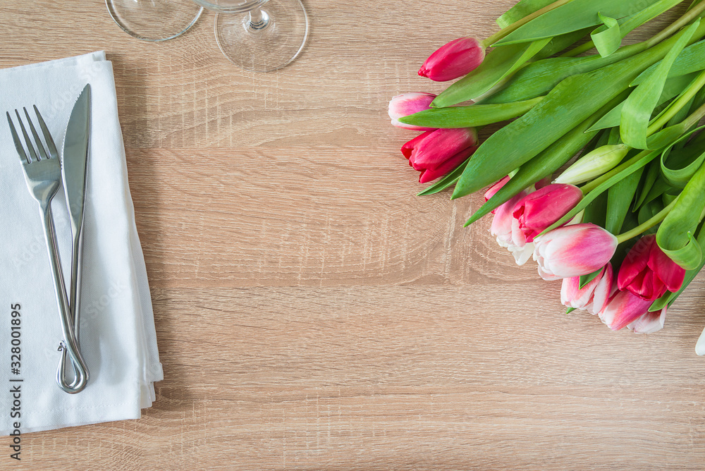 Festive table setting with bunch of purple tulips, forged fork and knife and set of wineglasses on white napkin and wooden background. Space for text, top view