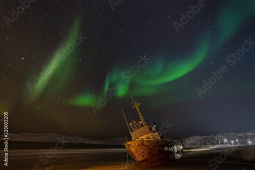 Polar lights in the starry sky. A fishing boat lying on its side, washed up by a storm on the Bank of the Barents sea. Teriberka, Russia