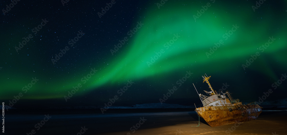 Naklejka premium Polar lights in the starry sky. A fishing boat lying on its side, washed up by a storm on the Bank of the Barents sea. Teriberka, Russia